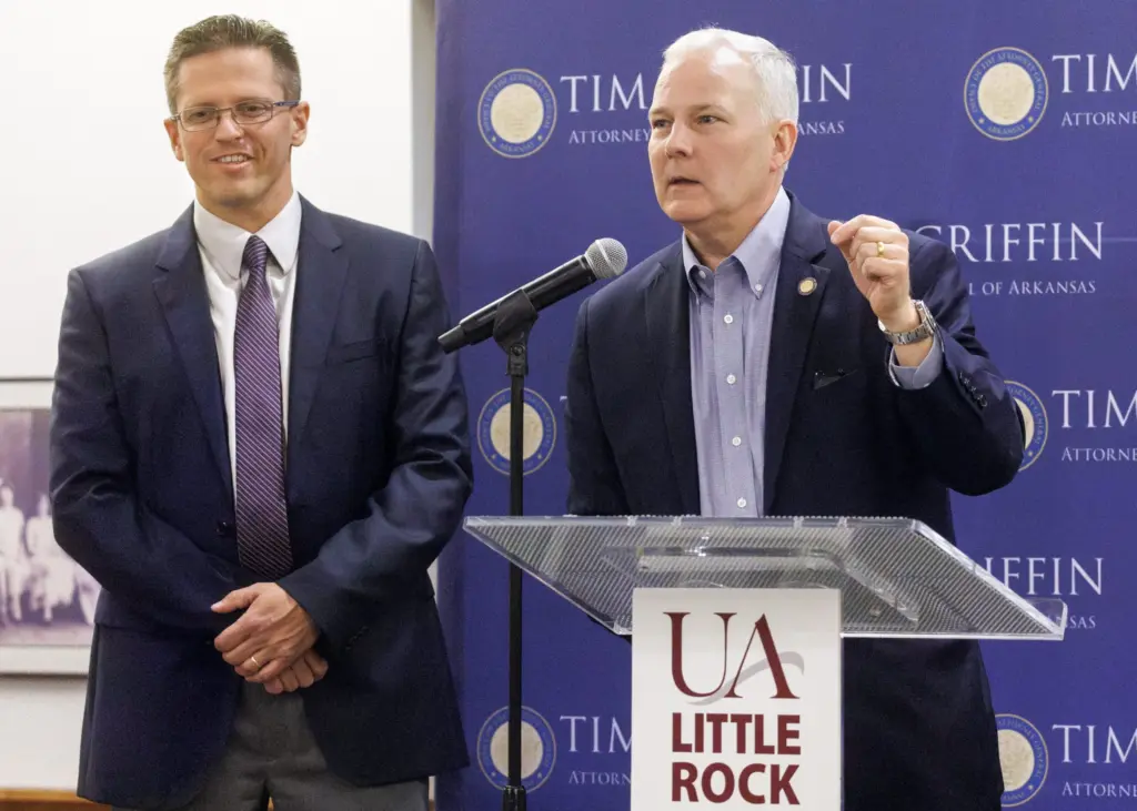 Two men in dark blue suits with one of the men talking into a microphone.