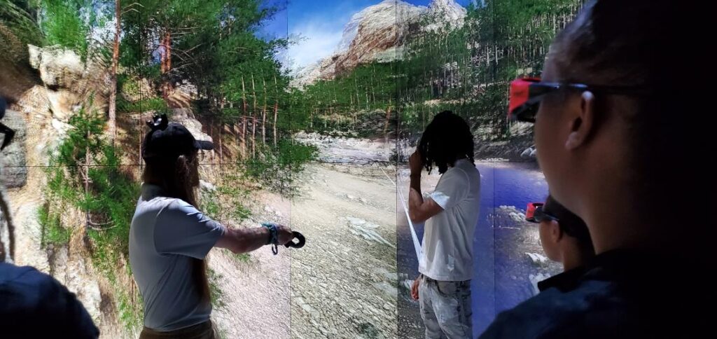 A group of students stand around a man pointing toward a screen that is displaying a digital forest.