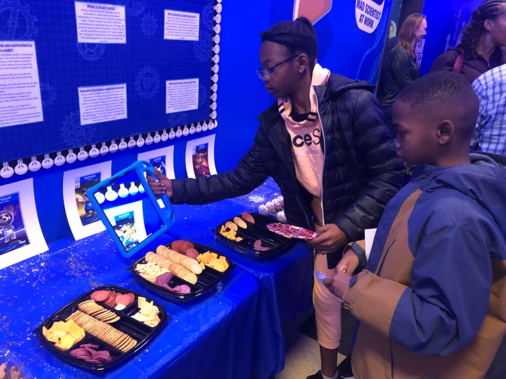 A girl holding a plate of snacks in her left hand holds a tablet in another pointed at a poster with a boy standing beside her.