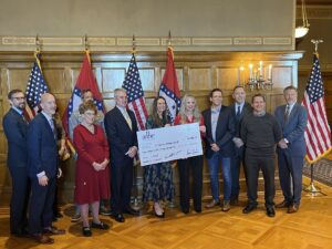 A group of men and women holding a large paper check in front of a line of flags.